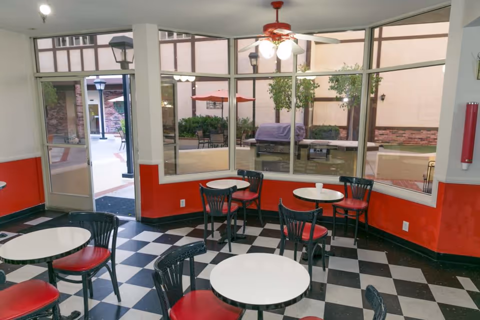 Indoor seating area with small round tables and black chairs with red cushions, black and white checkered floor, red and white walls, large windows looking out to an outdoor patio with tables, chairs, umbrellas, and greenery.