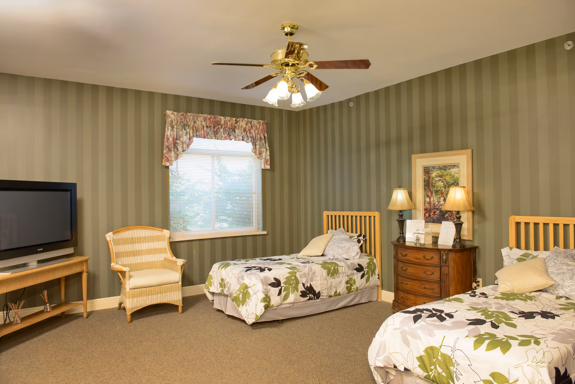 A bedroom with two twin beds featuring floral bedspreads, a wooden nightstand with two lamps and a framed picture, a wicker chair, a flat-screen TV on a wooden stand, a window with floral valance curtains, and a ceiling fan with lights.