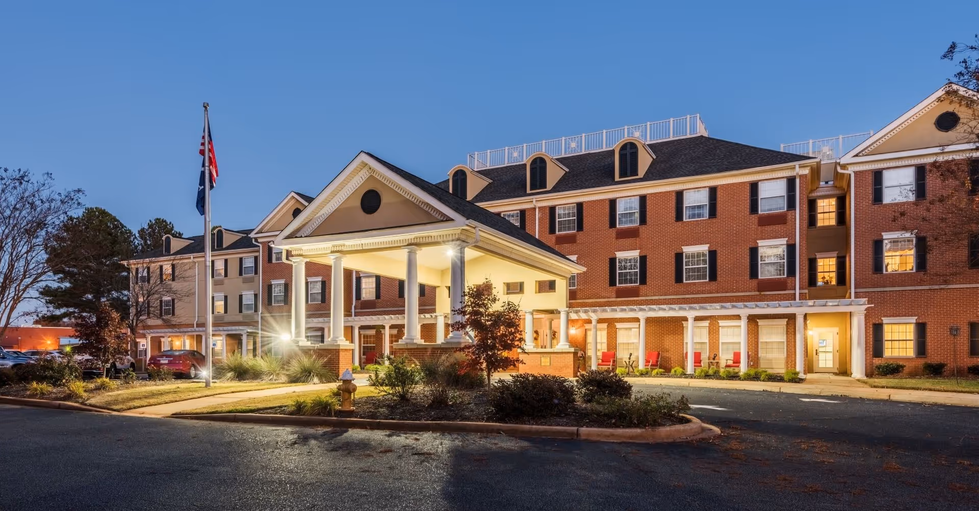 Exterior view of TerraBella Thornblade senior living facility at dusk, showing a large brick building with multiple windows, a covered entrance with columns, a flagpole with flags, and a landscaped area with bushes and a fire hydrant.