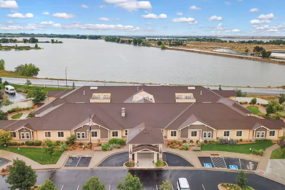 Aerial view of Seven Lakes Memory Care facility showing a single-story building with a brown roof and beige walls, surrounded by landscaped greenery and parking spaces including handicapped spots. Behind the building is a large body of water with a road running alongside it, under a partly cloudy sky.