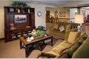 A cozy living room area in a senior care facility featuring two olive-green sofas with cushions, a wooden coffee table decorated with plants and tissues, and a large wooden entertainment center with a TV and decorative items. The room has warm lighting and a view into a hallway and dining area in the background.