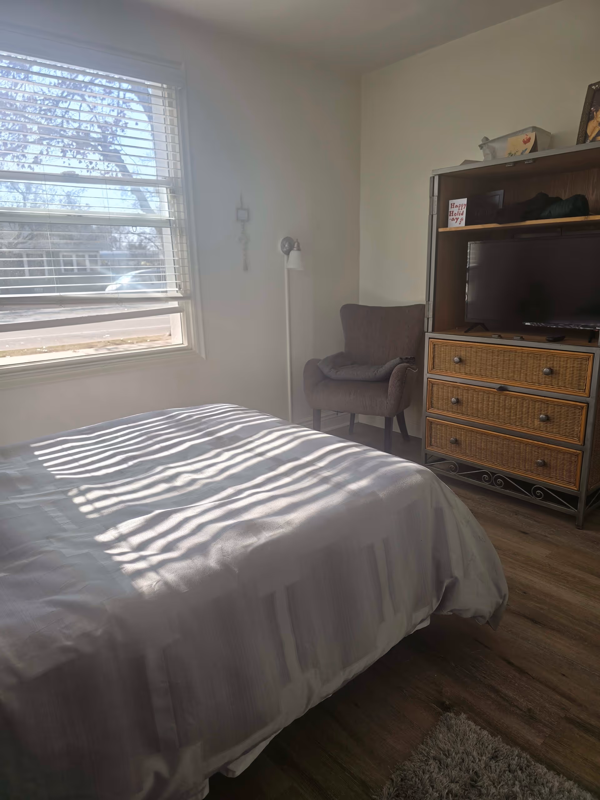 A sunlit bedroom featuring a bed with striped bedding, a chair, and a wicker-front dresser with a TV beside a window.