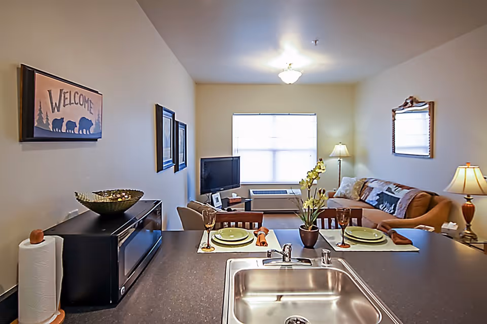 View from a kitchen counter with a sink looking into a living room area. The counter has two place settings with green plates, brown napkins, and glasses. A microwave and paper towel holder are on the left side of the counter. The living room has a couch with pillows, a floor lamp, a TV on a stand, and a window with blinds. Wall decorations include a 'Welcome' sign with bear silhouettes and framed pictures.