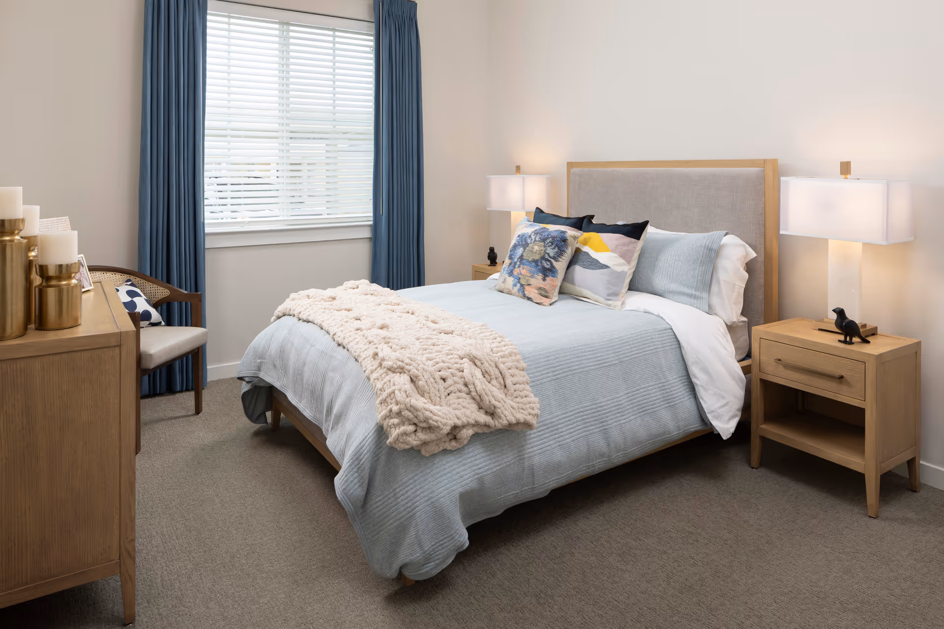 A cozy bedroom with a neatly made bed featuring light blue bedding, decorative pillows, and a chunky knit throw blanket. The room has beige carpet, a wooden nightstand with a lamp and small bird figurine, a wooden dresser with candles, and a chair with a cushion. Blue curtains frame a window with white blinds.