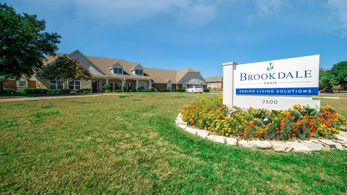 Exterior view of Brookdale Ennis senior living facility with a large lawn and a flower bed surrounding a sign that reads 'Brookdale Ennis Senior Living Solutions 2500' under a clear blue sky.