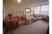A cozy living room area with beige carpet, a sofa, two armchairs, a coffee table, and a mirror on the wall. To the left, there is a small dining table covered with a red tablecloth and four wooden chairs. A large window with vertical blinds allows natural light into the room.
