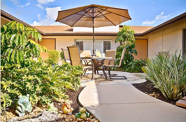 Outdoor patio area with a round table and four chairs under a large beige umbrella. The patio is surrounded by green plants and small garden statues of frogs and a squirrel. The sky is blue with some clouds.