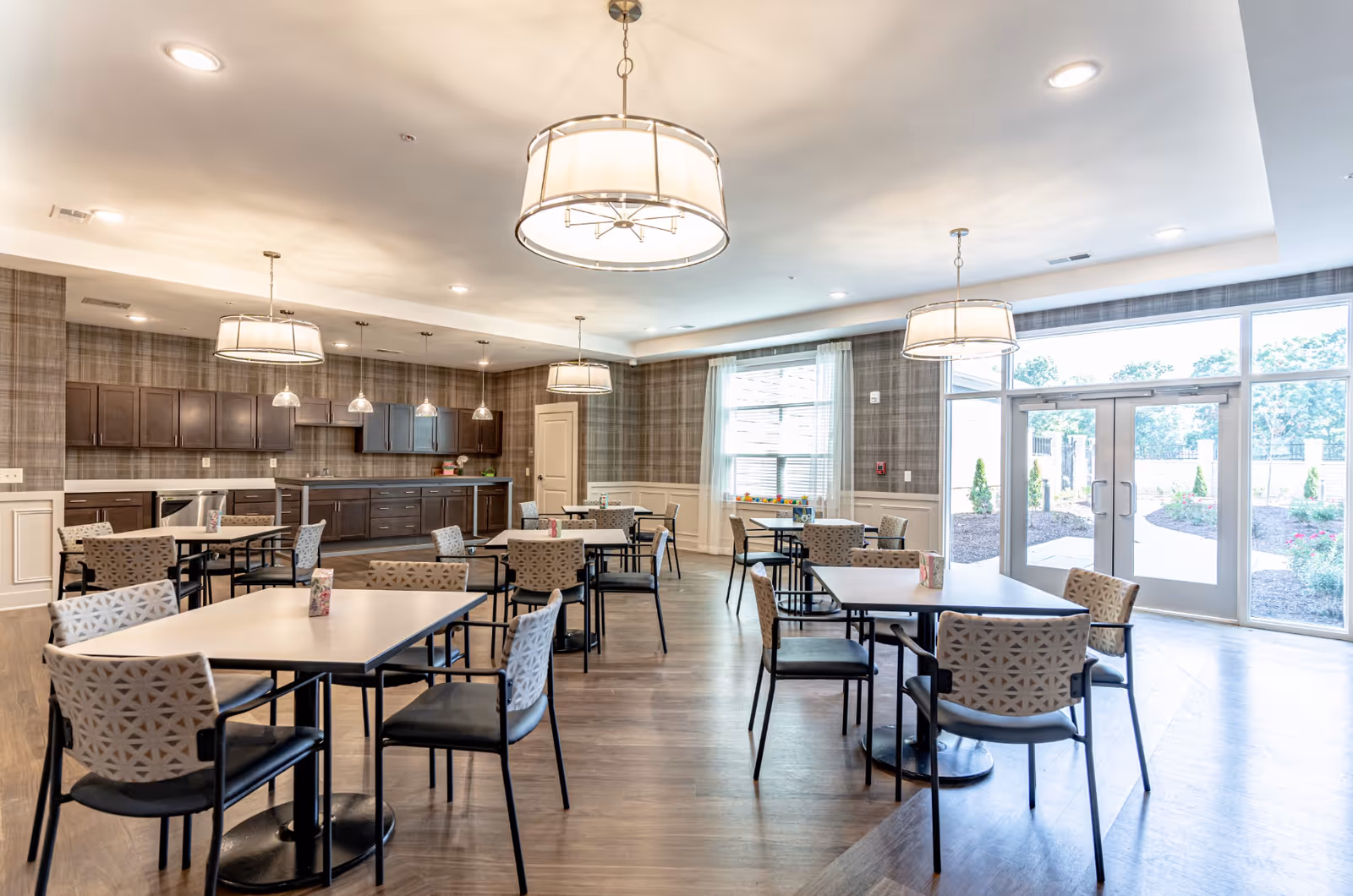 A bright dining room with multiple square tables and patterned chairs arranged neatly. The room features large windows and glass doors letting in natural light, modern pendant lights hanging from the ceiling, and a kitchen area with dark wood cabinets and a dishwasher in the background.