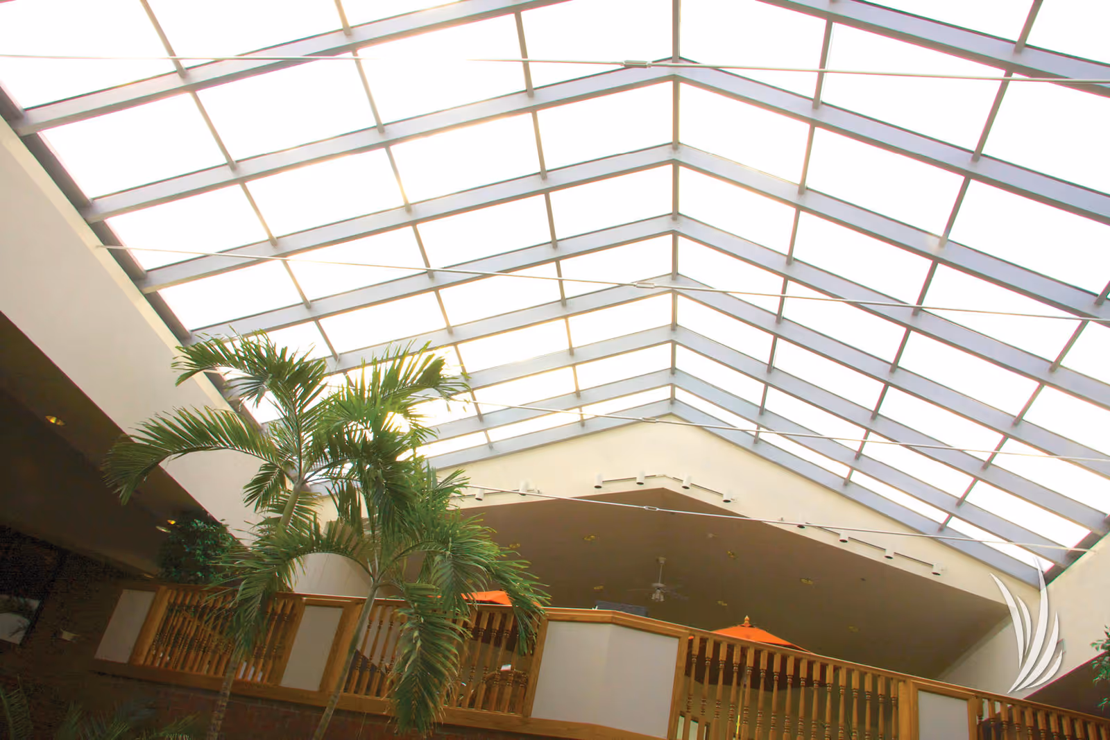 Interior view of a senior living facility with a large glass ceiling allowing natural light to fill the space. There are indoor palm plants and a wooden railing on an upper level with orange umbrellas visible in the background.