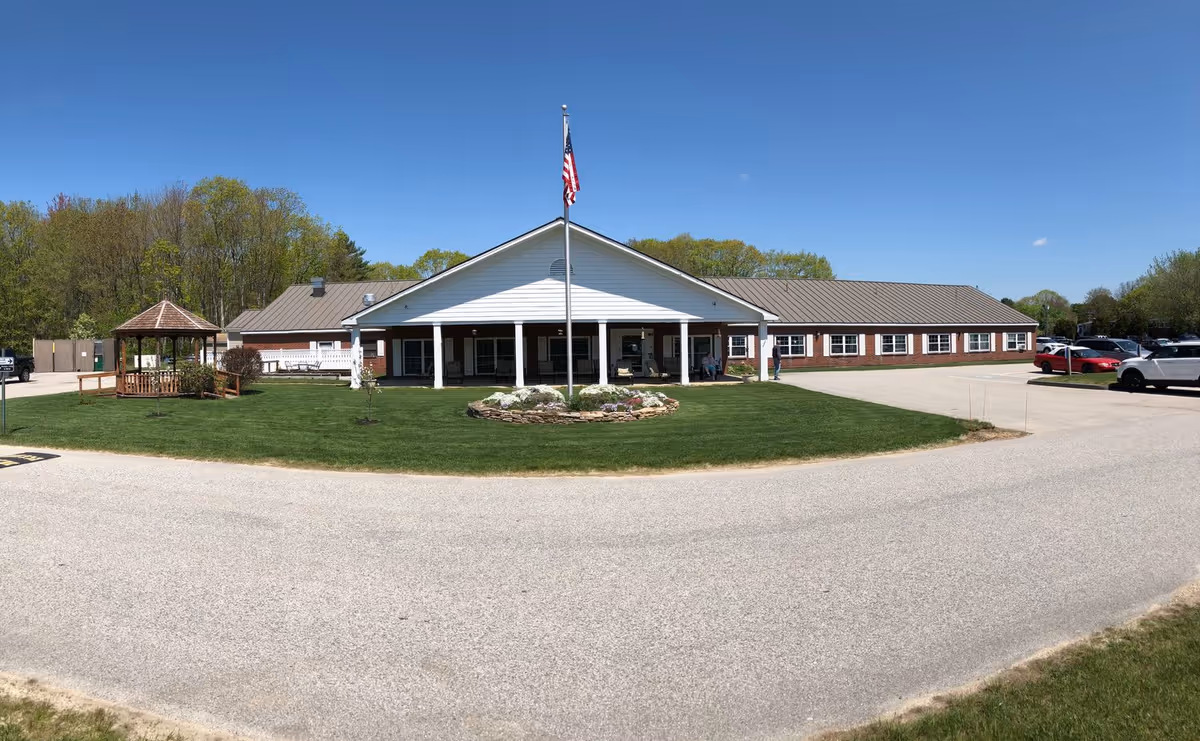 A single-story brick-and-white senior living building with a central covered entrance, flagpole, gazebo, lawn and parking under a clear blue sky.