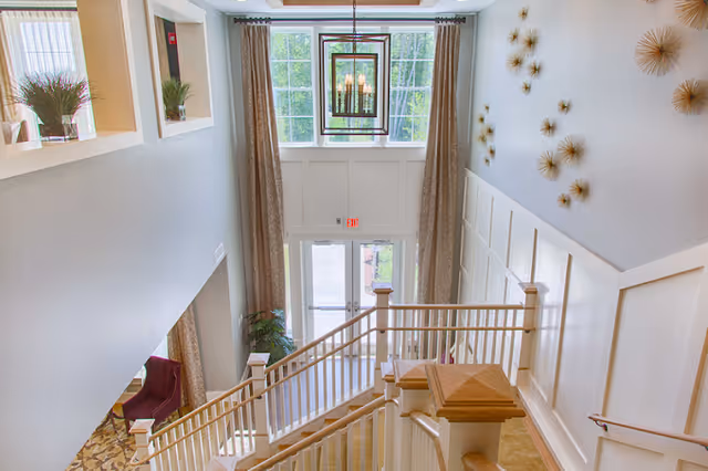 View from the top of a staircase looking down towards a glass door entrance with large windows above it. The walls are light-colored with decorative paneling and gold starburst wall art. There are beige curtains framing the windows and a modern chandelier hanging from the ceiling. A red armchair and plants are visible in the lower left corner.
