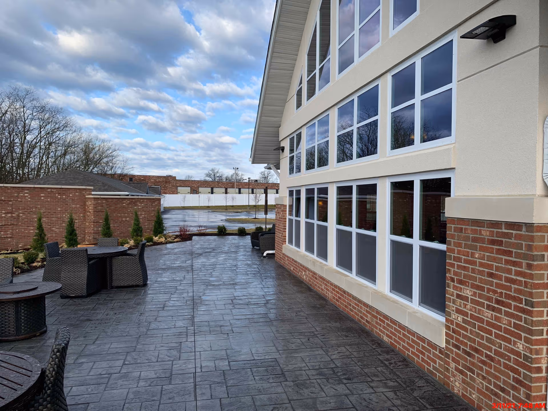 Outdoor patio area at Creek Valley Retirement Community with dark stone flooring, several round tables with wicker chairs, a brick and stucco building with large windows, small shrubs along a brick wall, and a cloudy sky overhead.