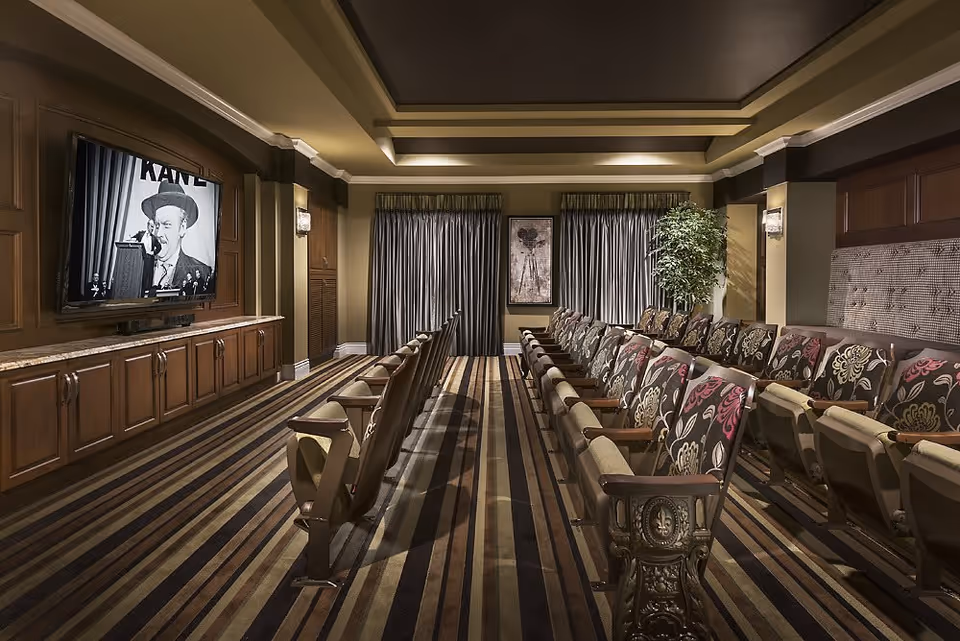 A cozy home theater room with two rows of upholstered chairs facing a large flat-screen TV mounted on a wooden paneled wall. The room features striped carpeting, soft lighting, and curtains covering the windows. A framed artwork and a potted plant are visible at the back of the room.