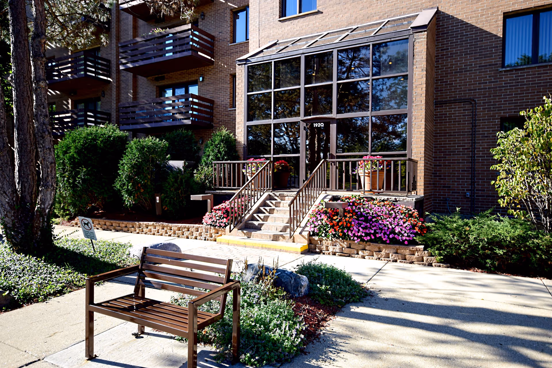 Exterior view of Valley Lo Towers II Apartments showing the entrance with large glass windows and a door numbered 1920. There are stairs leading up to the entrance, flanked by flower beds with colorful flowers. A wooden bench is placed on the sidewalk in front of the building, surrounded by greenery and trees.