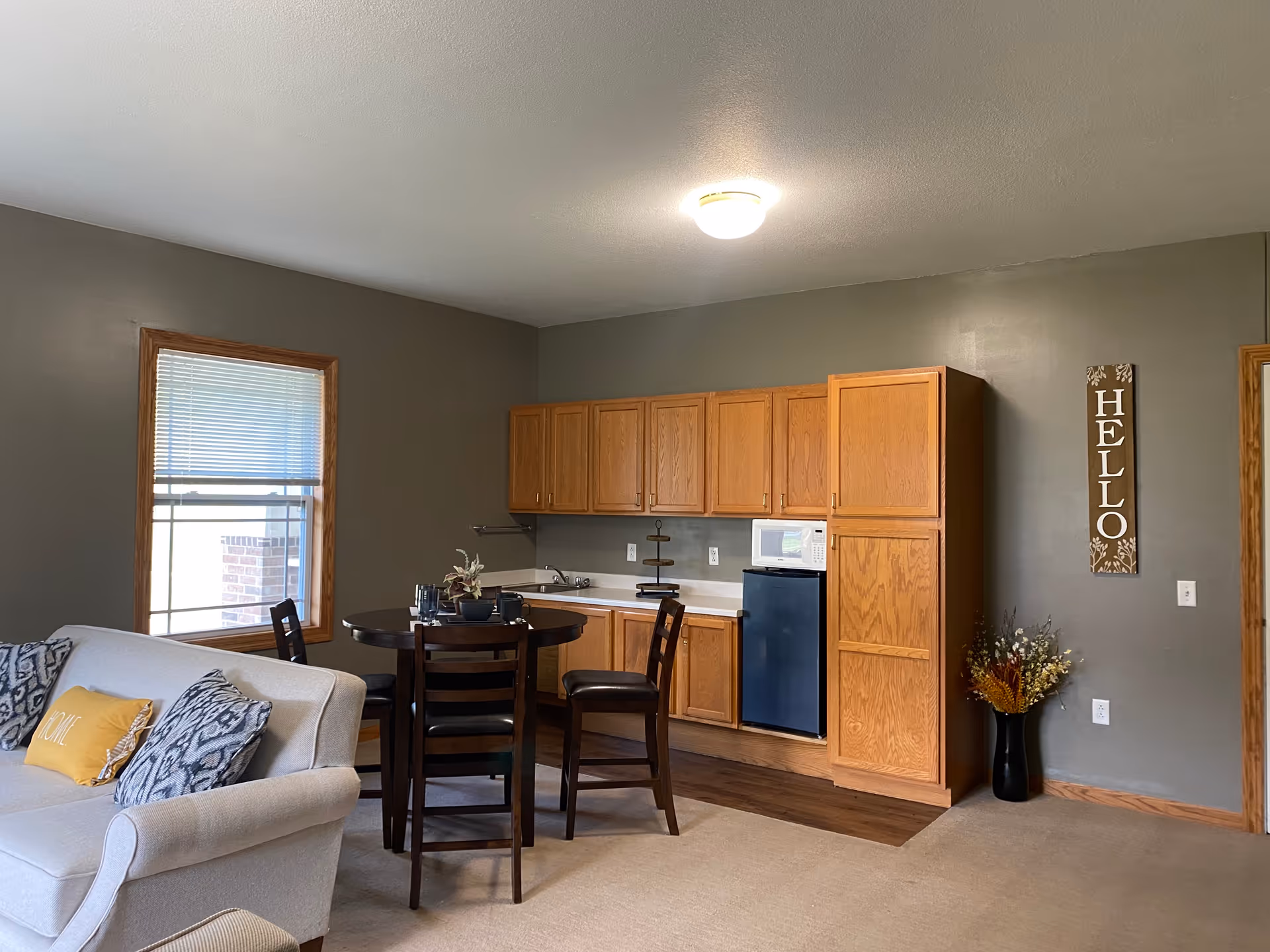 A cozy living area with a light gray couch adorned with patterned and yellow pillows, a round dark wooden dining table with four chairs, and a kitchenette featuring wooden cabinets, a small refrigerator, a microwave, and a sink. A window with blinds is on the left wall, and a decorative 'HELLO' sign hangs on the right wall above a vase with dried flowers.