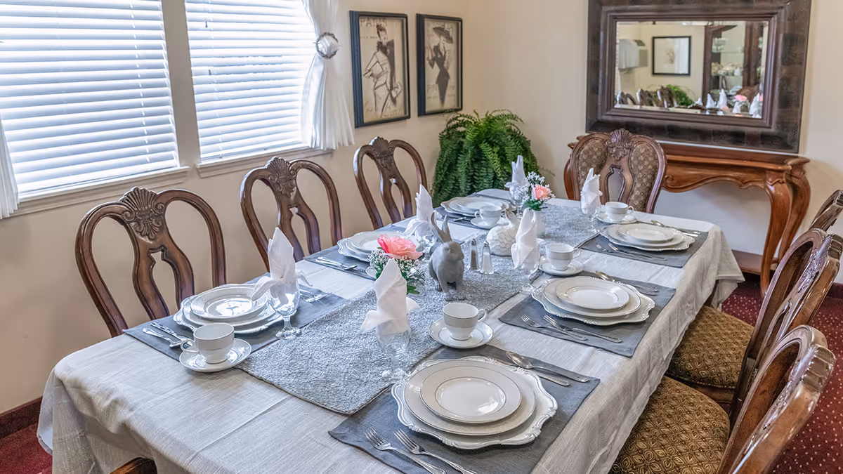 Formally set dining table with place settings, napkins, and wooden chairs in a bright dining room.