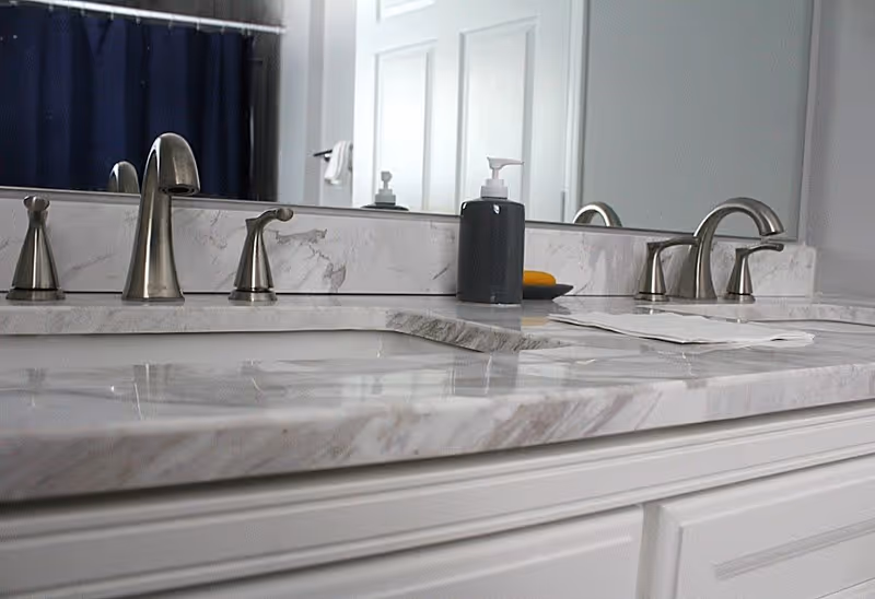 Close-up view of a bathroom countertop with a marble surface, featuring two silver faucets, a soap dispenser, and a mirror reflecting a shower curtain and a door.