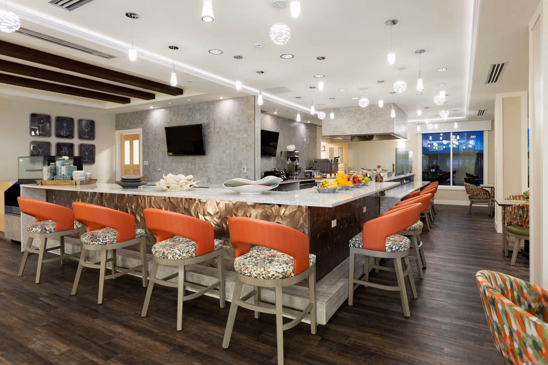 Bright communal dining area with a long marble-topped island surrounded by orange-cushioned bar stools and an open kitchen in the background.