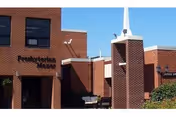 Brick-front entrance of a Presbyterian Manors facility with a small courtyard, bench, and a tower-like steeple.