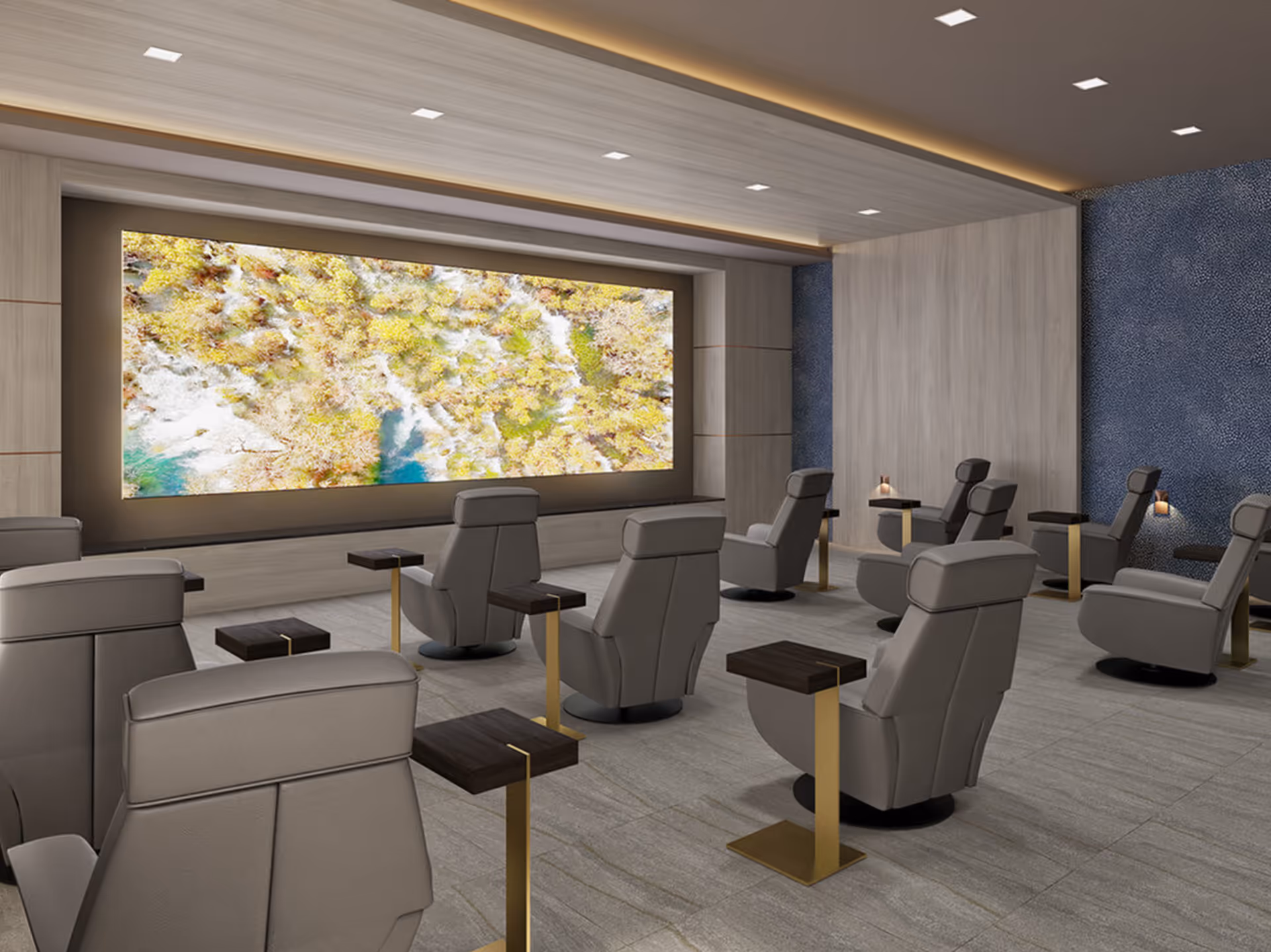 A modern media room with multiple gray reclining chairs arranged in rows facing a large screen displaying an aerial view of a forest and river. Each chair has a small side table with a dark wood top and gold base. The room features light wood paneling on the walls and ceiling, with recessed lighting and a textured blue accent wall on the right.