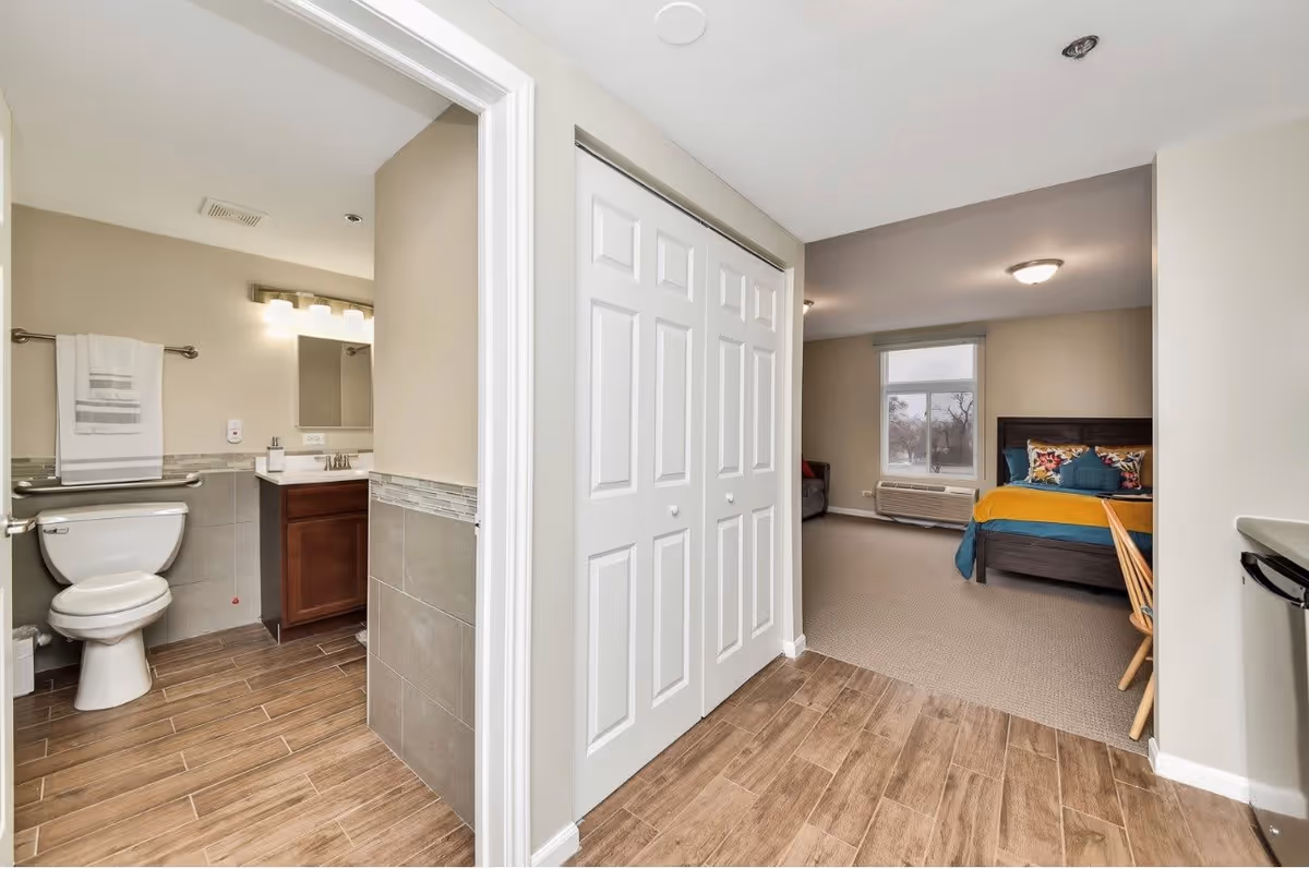 View of an interior space in a senior living facility showing a bathroom with a toilet, sink, mirror, and towel rack on the left, and a bedroom area with a bed, colorful pillows, a window, and a chair on the right. The floor transitions from wood-like tile in the hallway and bathroom to carpet in the bedroom.