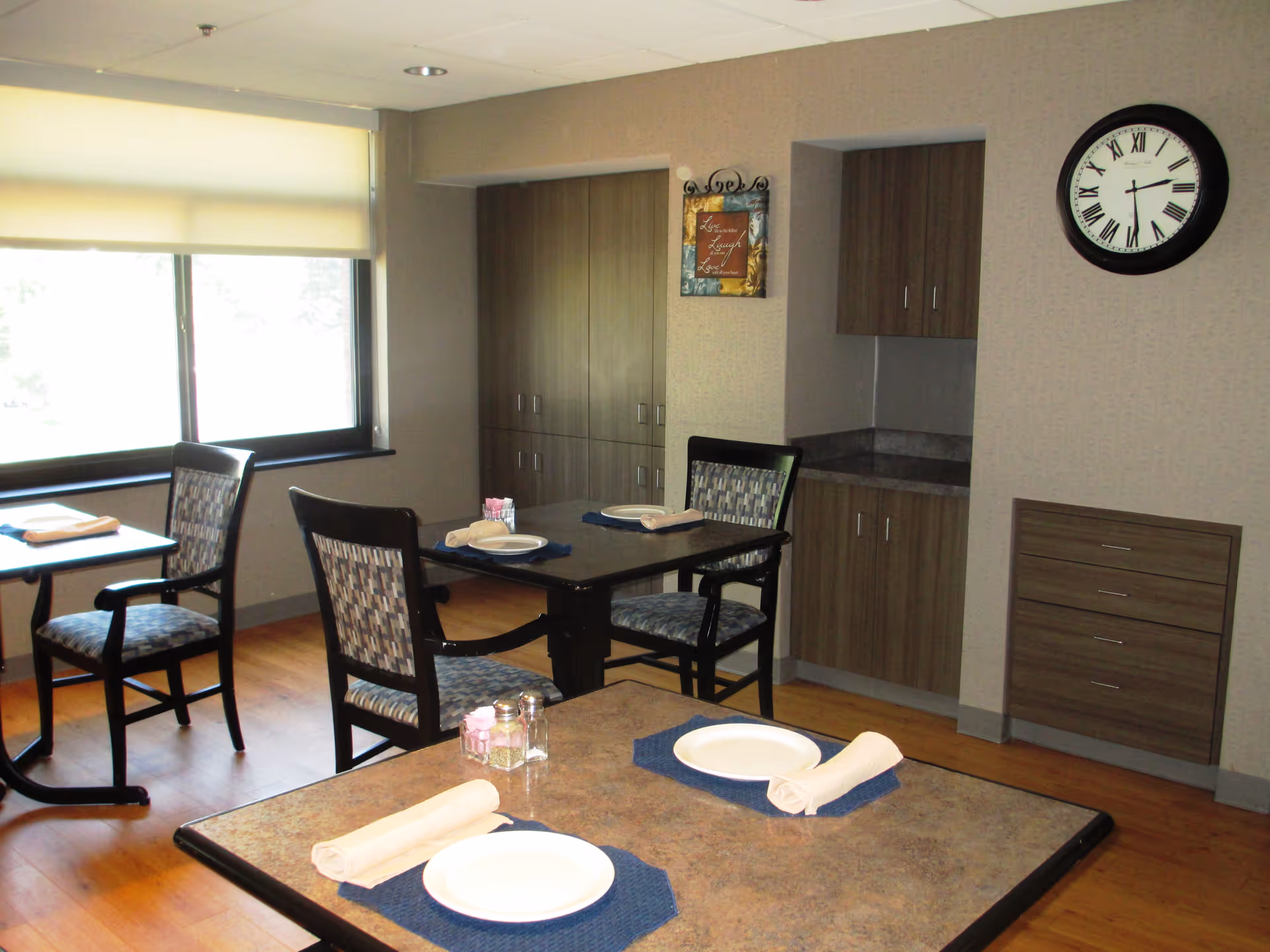 A dining area with several tables and chairs set for meals. Each table has white plates, rolled napkins on blue placemats, and salt and pepper shakers. The room has wooden flooring, a large window with a roller shade, built-in cabinets, and a wall clock showing the time as 2:10.