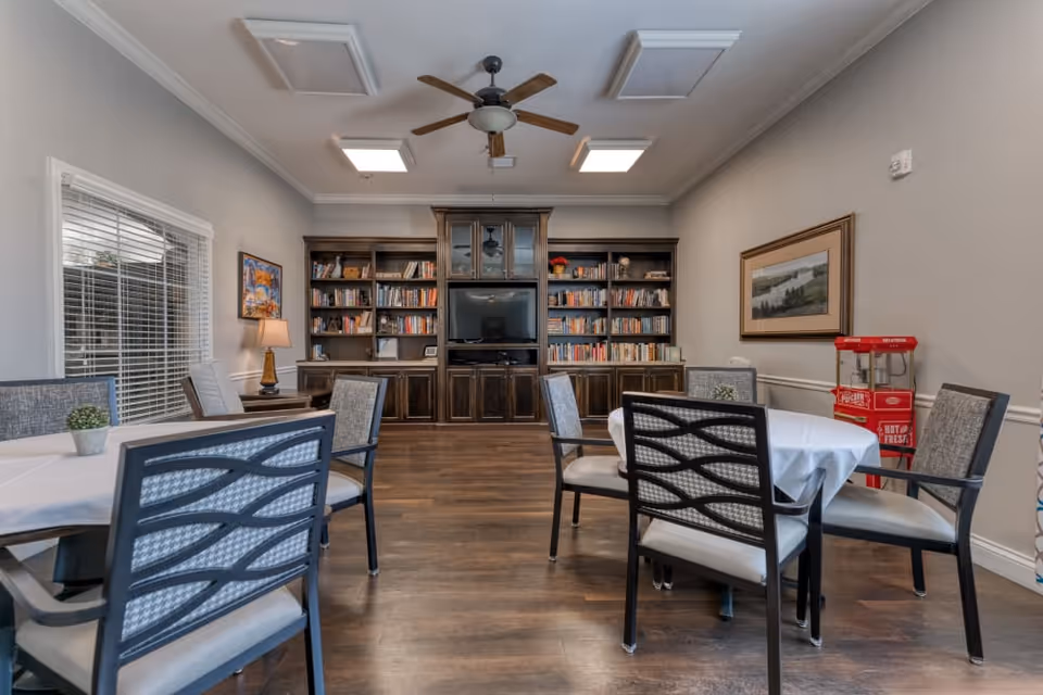 A cozy common area with round tables covered with white tablecloths and surrounded by cushioned chairs. The room features a large wooden bookshelf filled with books and a television in the center. A ceiling fan with wooden blades is mounted above, and a red popcorn machine is visible in the corner. The walls are decorated with framed artwork, and a window with blinds lets in natural light.