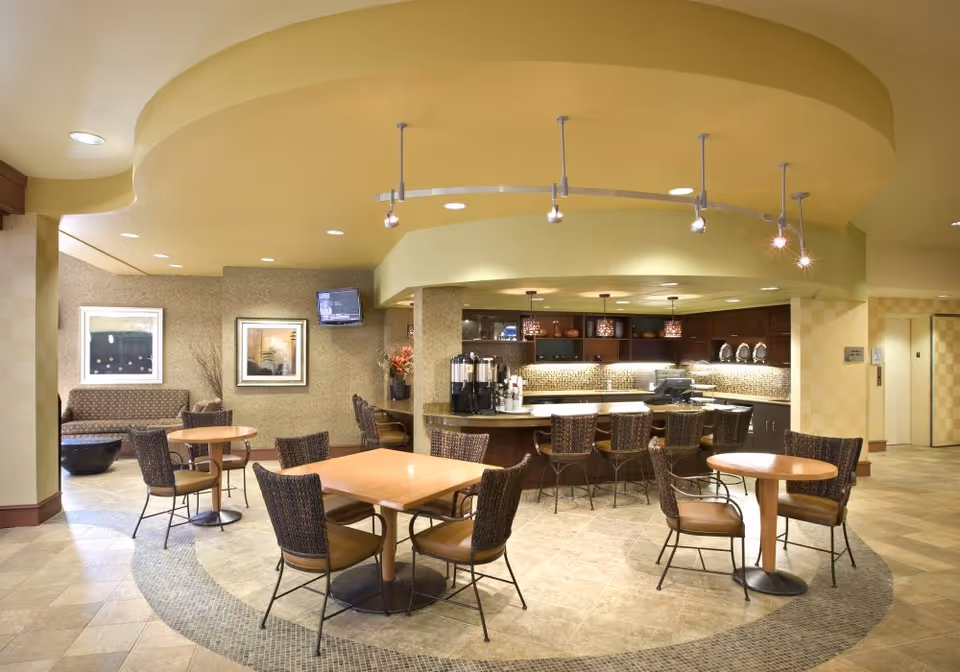 A well-lit common area with a coffee bar and seating at Fairwinds - Desert Point. The space features several round and square tables with wicker chairs, a curved ceiling design with track lighting, a coffee station with dispensers, and a small lounge area with a couch and framed artwork on the walls.