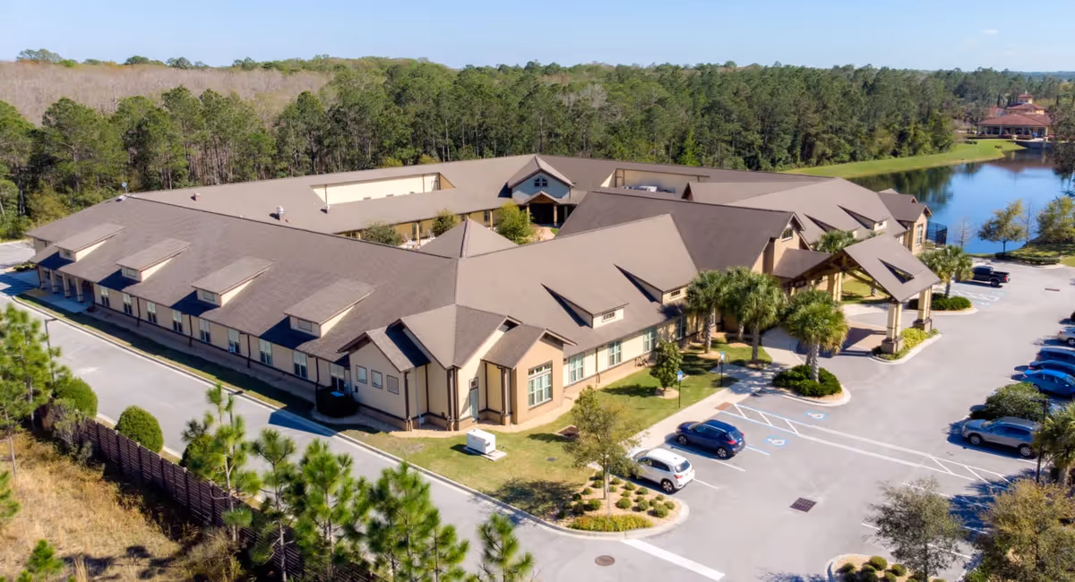 Aerial view of Rosecastle at Deerwood, a large single-story senior living facility with a brown roof, surrounded by parking spaces, trees, and a pond in the background.