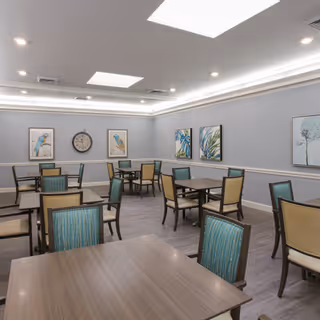 A well-lit dining room with multiple square wooden tables and chairs featuring green and beige upholstery. The walls are painted light gray with white trim and decorated with framed artwork, including images of birds and plants. A large clock is mounted on one wall. The ceiling has recessed lighting and skylights.