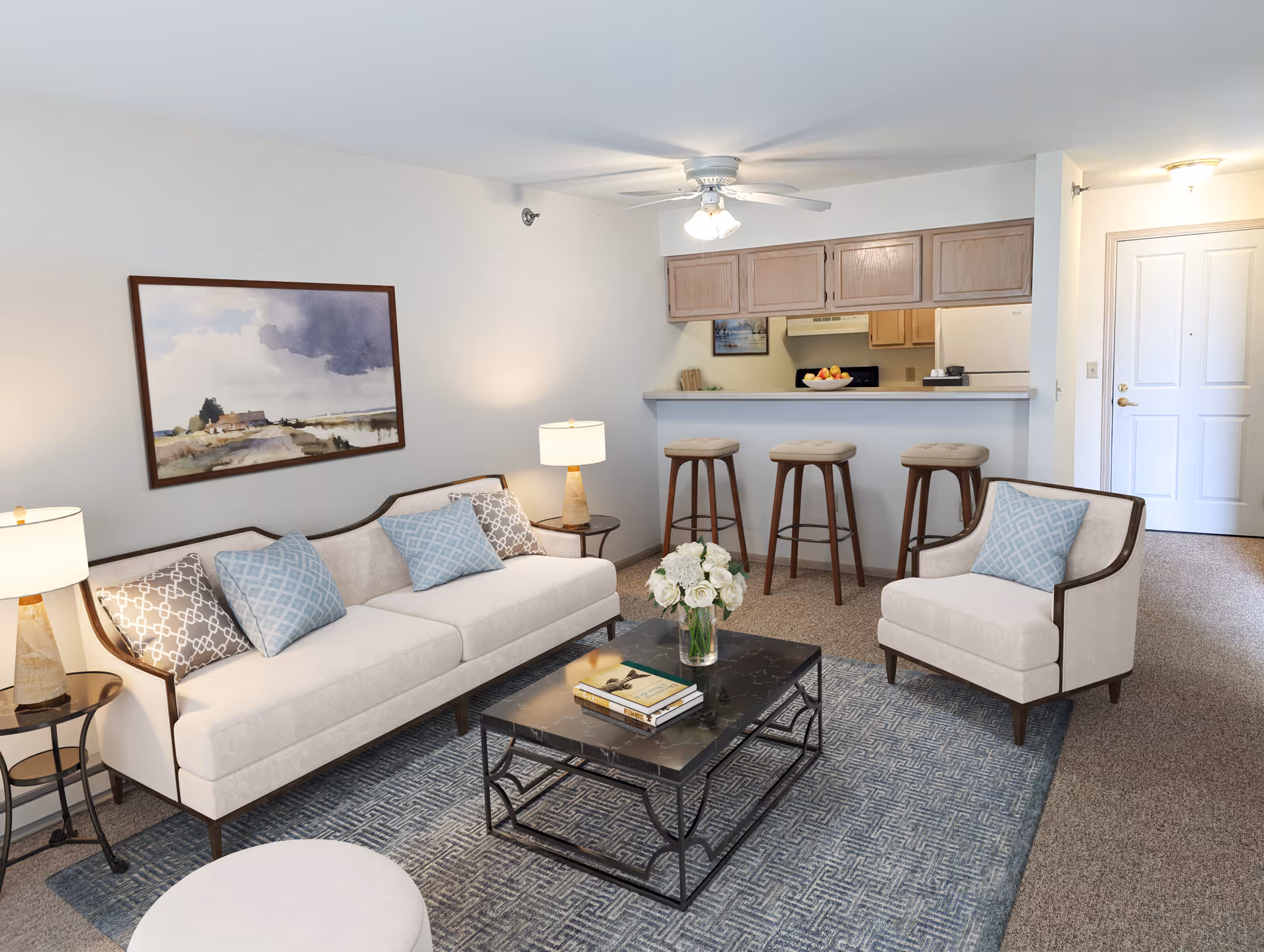 Bright furnished living room with a sofa, armchair, coffee table, area rug, and a breakfast bar with three stools opening to a kitchen.