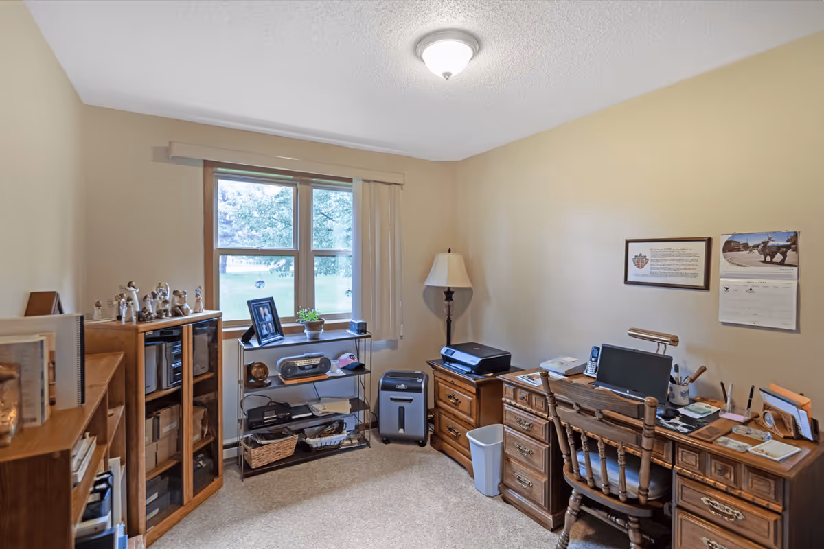 A small home office with beige walls and carpeted floor. There is a wooden desk with a computer, lamp, phone, and office supplies. Next to the desk is a wooden filing cabinet with a printer on top. A wooden chair is placed in front of the desk. Against the wall under a window is a metal shelving unit holding a radio, framed photo, and other items. A tall floor lamp stands in the corner. A wooden cabinet with glass doors and various figurines is on the left side of the room. The window shows a green outdoor area with trees.