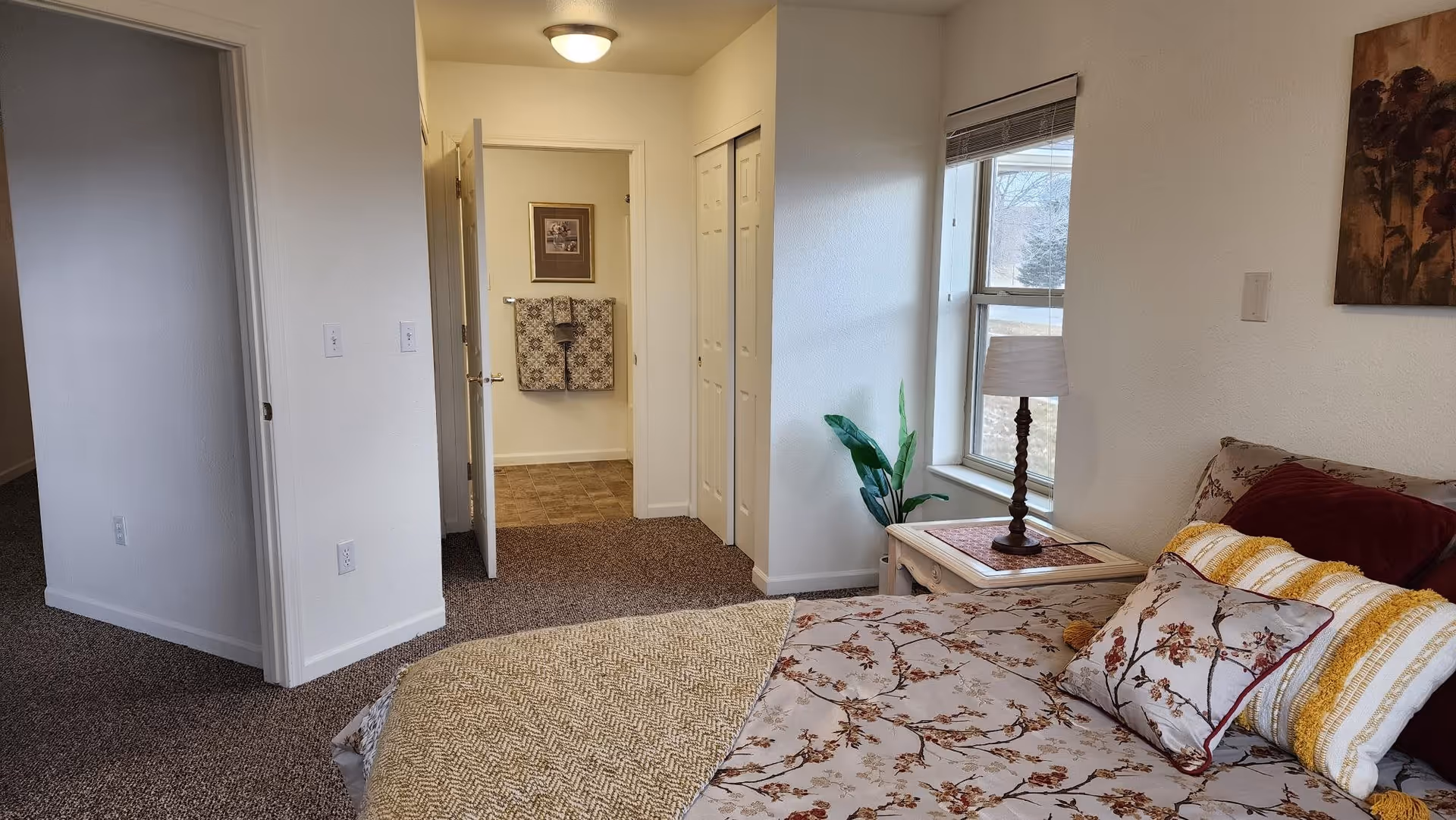 A bedroom with a bed covered in floral and striped pillows and a beige blanket. Next to the bed is a white nightstand with a lamp and a small plant. There is a window with blinds letting in natural light. The room has carpeted floors and white walls. In the background, an open door reveals a bathroom with a towel rack holding two patterned towels and a framed picture above it.