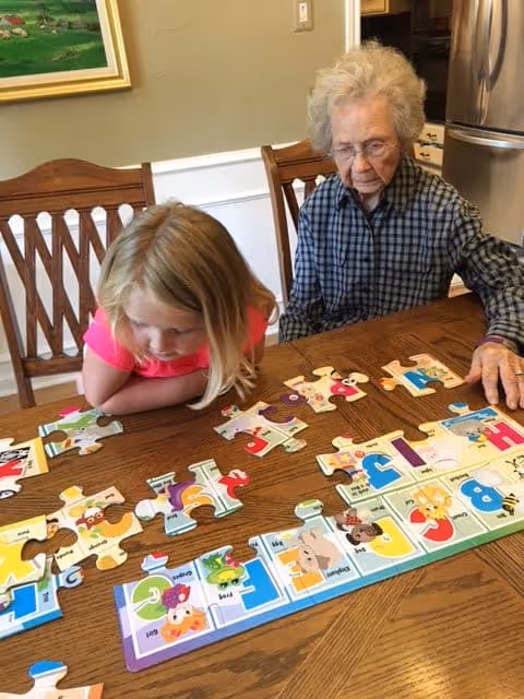 An elderly woman and a young girl sitting at a wooden table working together on assembling a colorful alphabet-themed jigsaw puzzle in a kitchen or dining area.