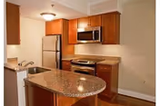 Interior view of a kitchen featuring wooden cabinets, a granite countertop island with a sink, a stainless steel refrigerator, microwave, and oven, with hardwood flooring and beige walls.