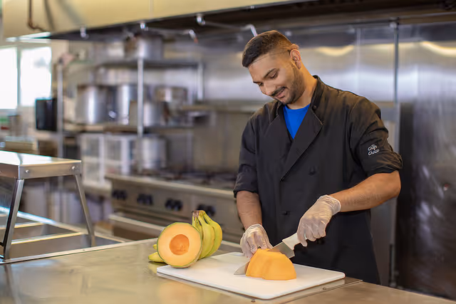 A man wearing a black chef coat and gloves is slicing a cantaloupe on a white cutting board in a commercial kitchen. There are bananas and half a cantaloupe on the counter beside him, with stainless steel kitchen equipment in the background.