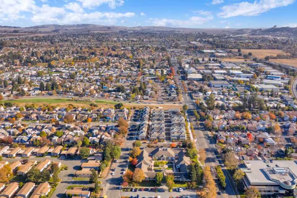Aerial view of a suburban area with numerous houses, buildings, and streets extending into the distance under a partly cloudy sky.