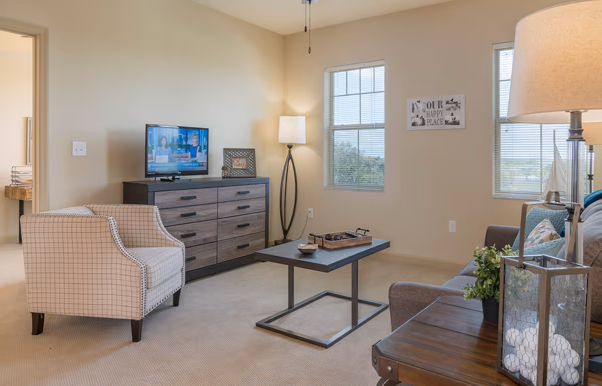 Bright furnished living room with an armchair, sofa, TV on a dresser, coffee table, and floor lamp near two windows.