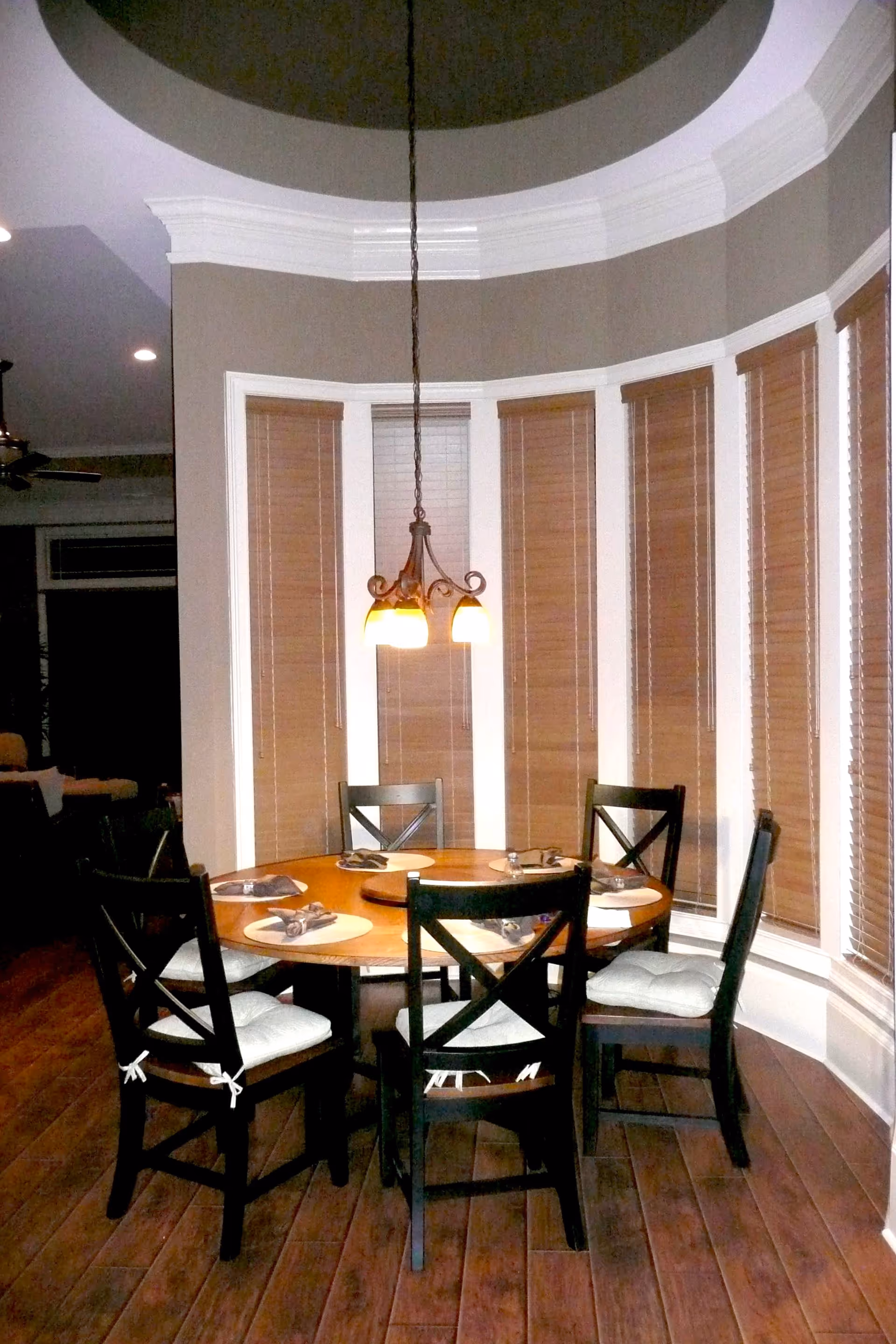 A round wooden dining table set with six black chairs with white cushions in a room with tall windows covered by brown blinds. A hanging light fixture with three lamps is above the table, which is set with placemats and napkins.