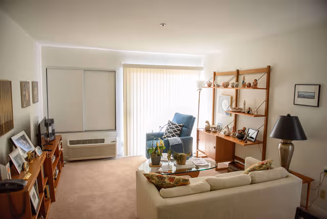 A cozy living room in a senior community featuring a beige sofa with patterned cushions, a glass coffee table with potted plants, a blue armchair with a decorative pillow, a wooden shelving unit with various decorative items, a floor lamp, and framed pictures on the walls. The room has carpeted flooring and vertical blinds covering a large window or sliding door.