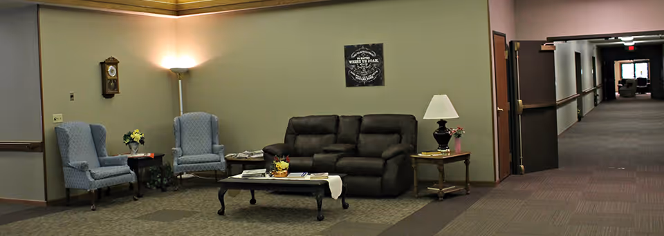 Interior view of a senior living facility lounge area with two blue armchairs, a dark brown sofa, a coffee table with a flower arrangement, a side table with a lamp and flowers, a wall clock, and a hallway leading to another room.