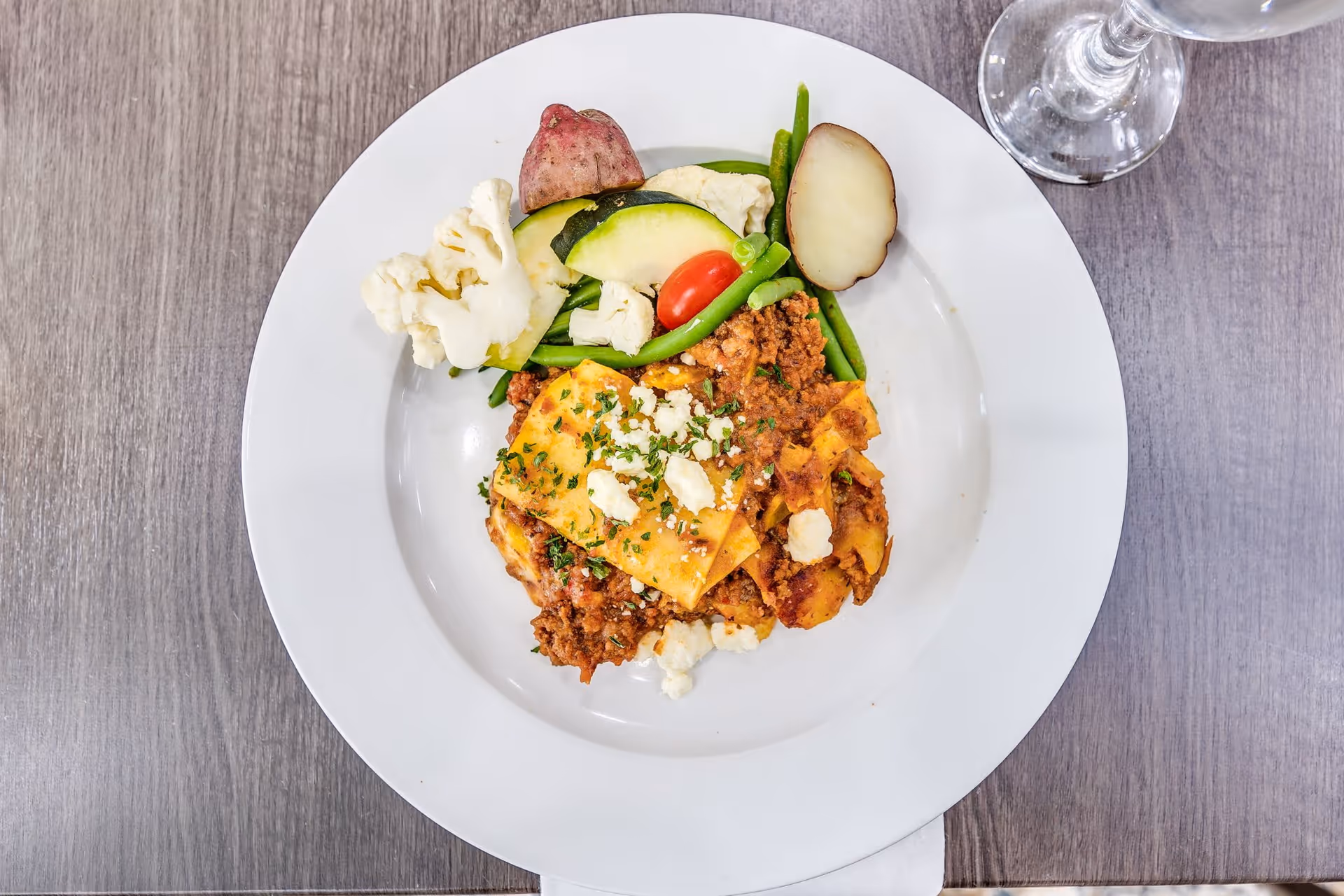 A white plate with a serving of lasagna topped with cheese and herbs, accompanied by steamed vegetables including cauliflower, green beans, zucchini, cherry tomato, and red potato slices, placed on a wooden table with a glass of water nearby.