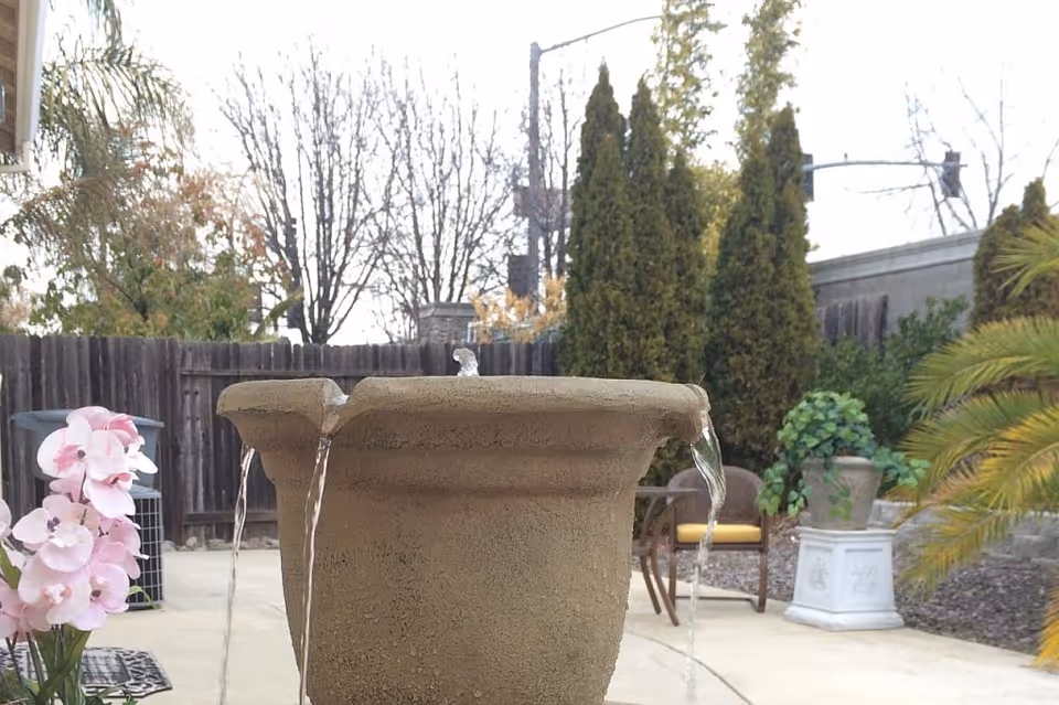 Outdoor patio area with a large stone water fountain in the foreground, water flowing from its edges. Surrounding the fountain are potted plants, including pink flowers and greenery. In the background, there are tall evergreen trees, a wooden fence, and patio furniture with a yellow cushion on one chair.