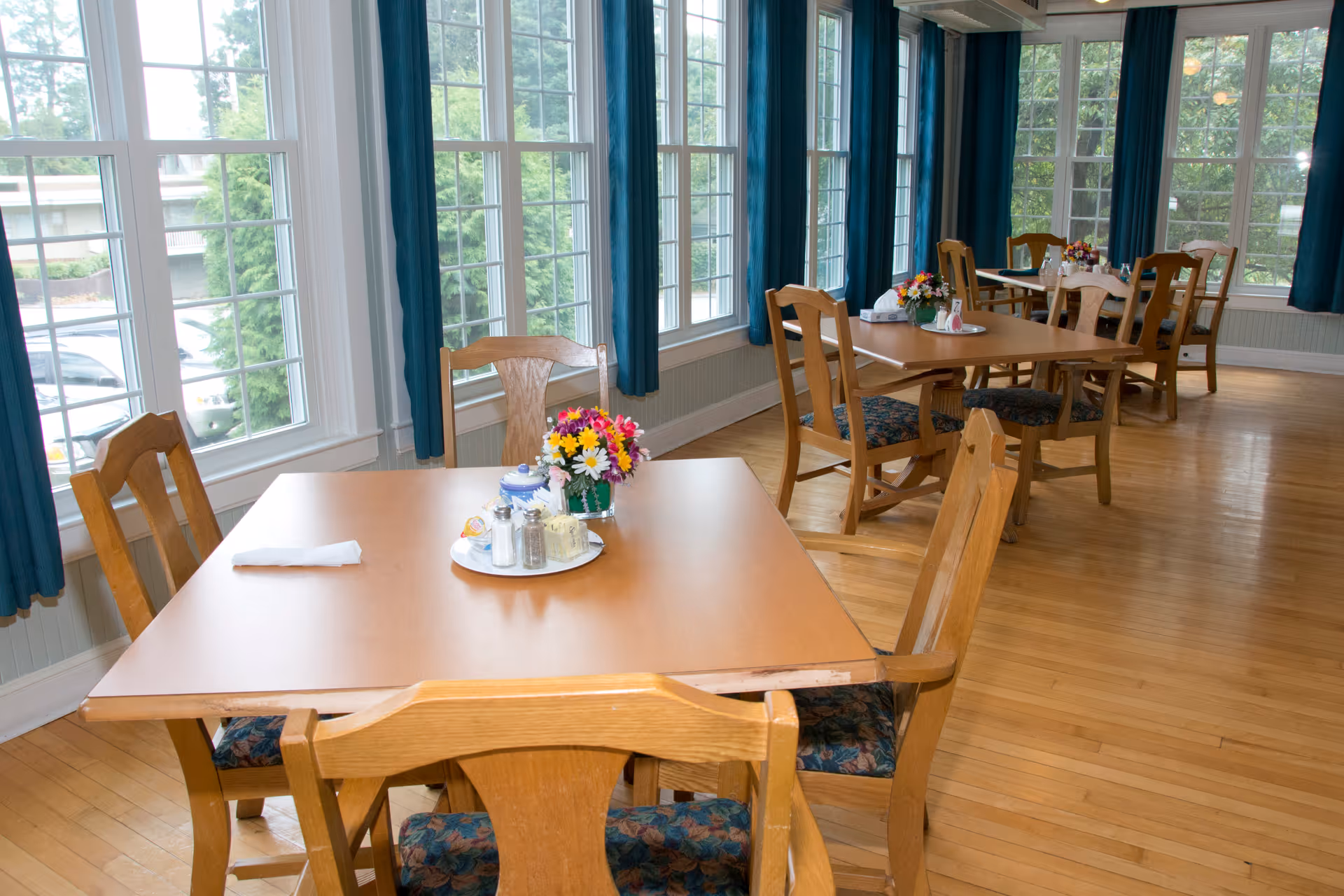 A bright dining room with wooden tables and chairs arranged neatly. Each table has a centerpiece with flowers and condiments. Large windows with blue curtains allow natural light to fill the room, and a wooden floor complements the warm atmosphere.
