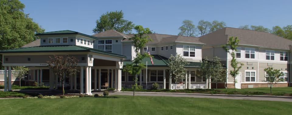 Exterior view of a two-story senior living facility building with beige siding and a green roof over the entrance. The building is surrounded by green grass, small trees, and shrubs under a clear blue sky.