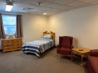 A simple bedroom with a single bed covered with a striped blanket, a wooden dresser under a window with curtains, a red upholstered armchair, and a small wooden side table with a remote control on it.