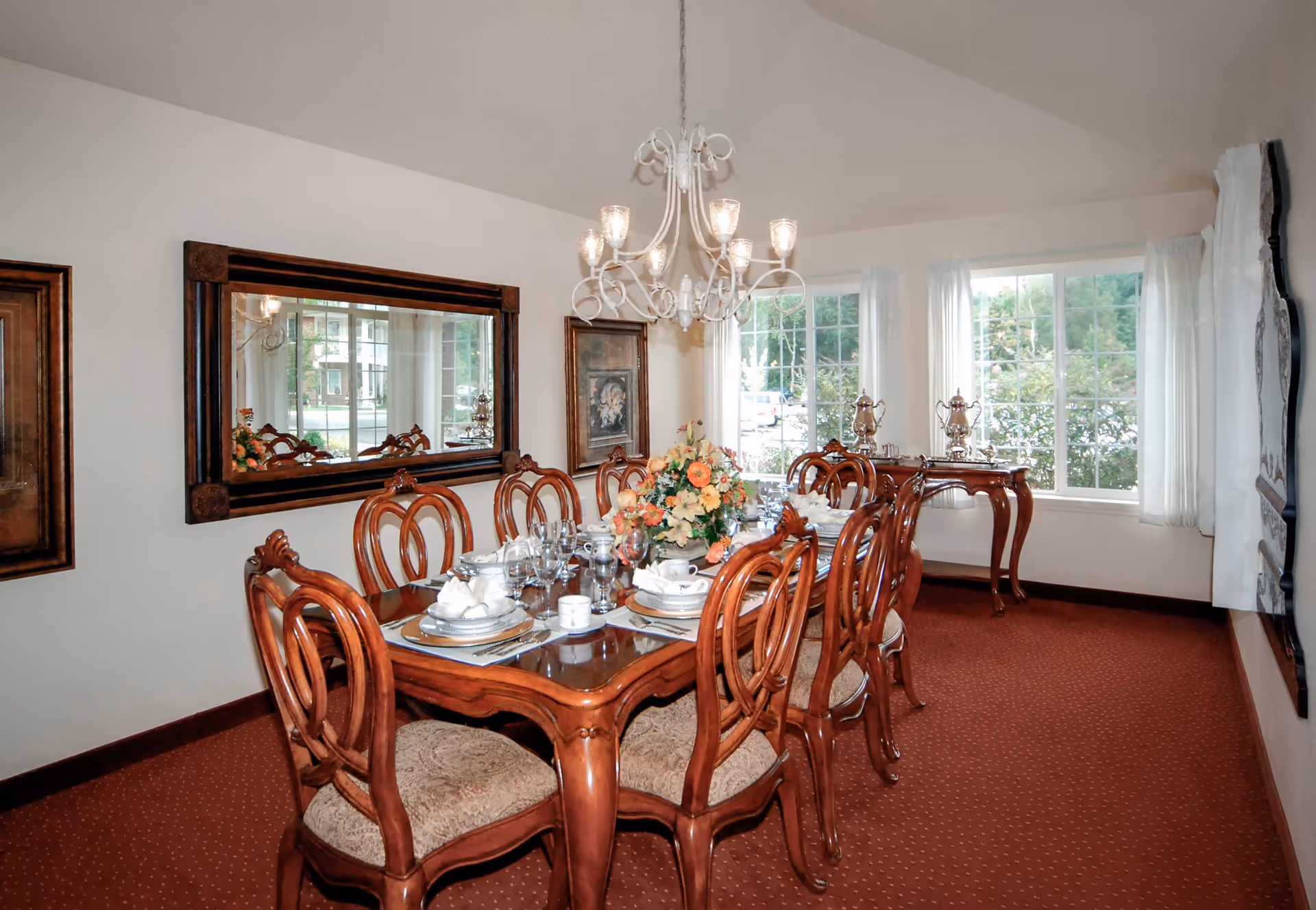 A formal dining room with a wooden dining table set for eight, featuring cushioned chairs, a floral centerpiece, and a chandelier overhead. The room has large windows with white curtains allowing natural light to enter, a large mirror on one wall, and a side table with silver tea sets.