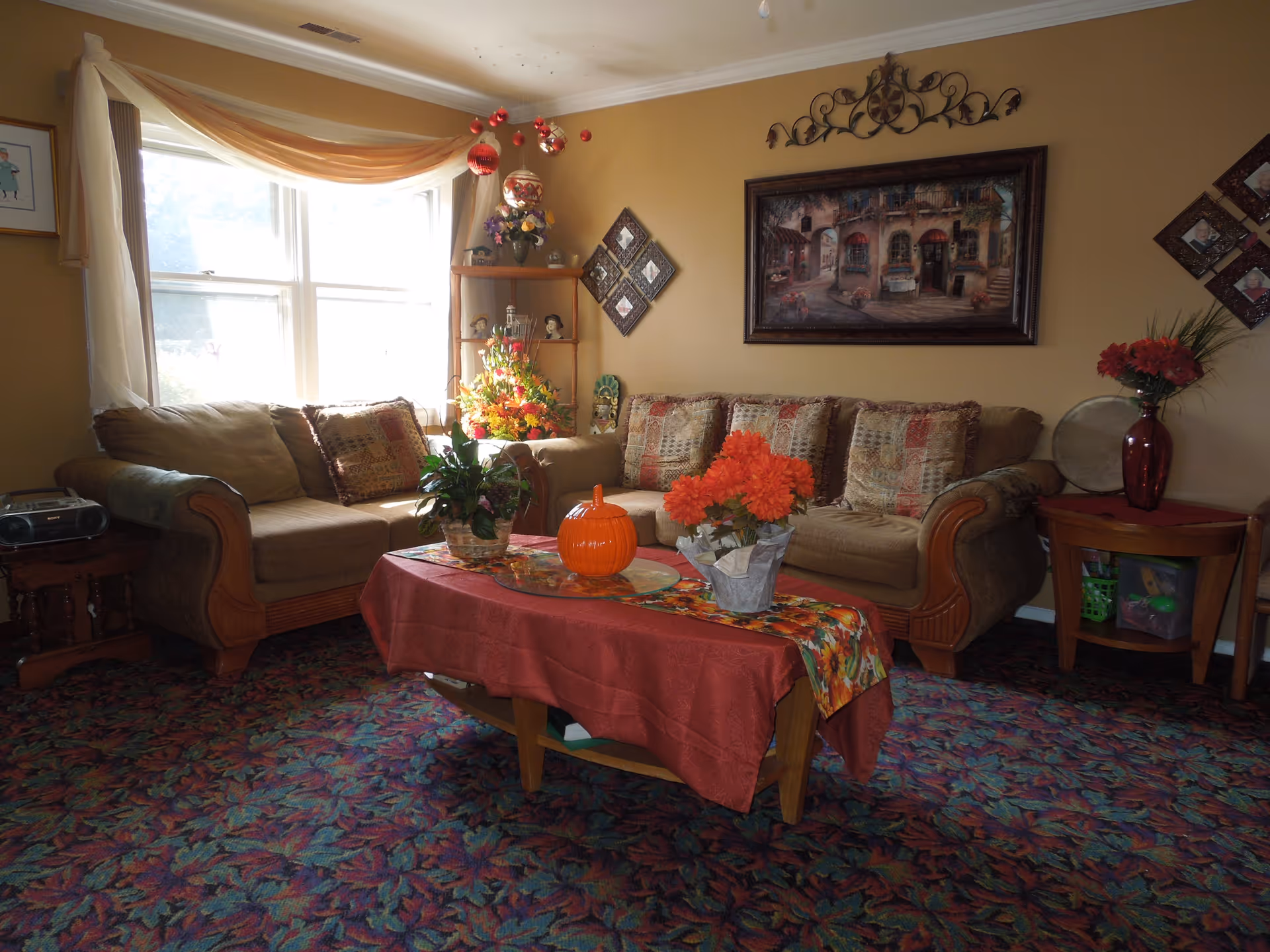 A cozy living room with two beige sofas adorned with patterned cushions. A wooden coffee table covered with a red tablecloth and a floral runner holds a small orange pumpkin decoration and two flower arrangements. The room features a large window with sheer curtains, a corner shelf with decorative items, and framed artwork on the walls. The carpet has a colorful leaf pattern.