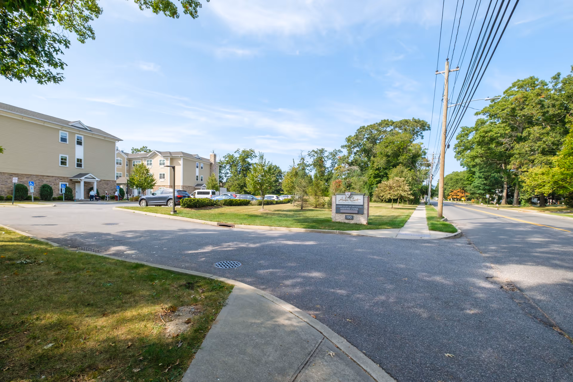 Exterior view of Amber Court assisted living facility showing a parking lot with cars, a sign with the facility name, and a three-story beige building with stone accents under a blue sky with some clouds.