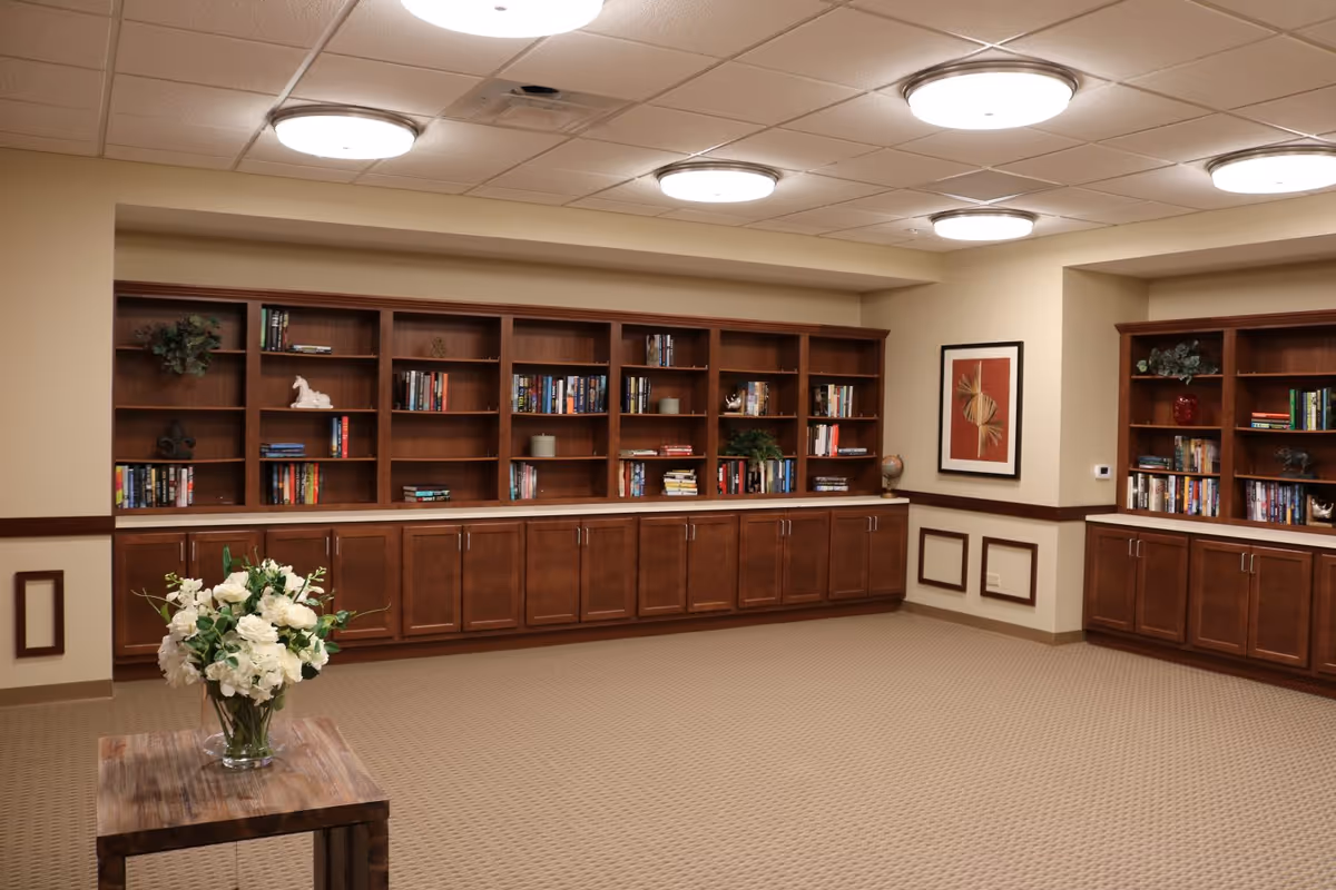 A spacious room with beige walls and carpeted floor featuring built-in wooden bookshelves filled with books and decorative items. A small wooden table with a vase of white flowers is in the foreground. The ceiling has multiple round light fixtures, and a framed abstract artwork hangs on the wall.